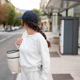 Person wearing a white sweatshirt and blue cap, holding a tote bag on a city street.