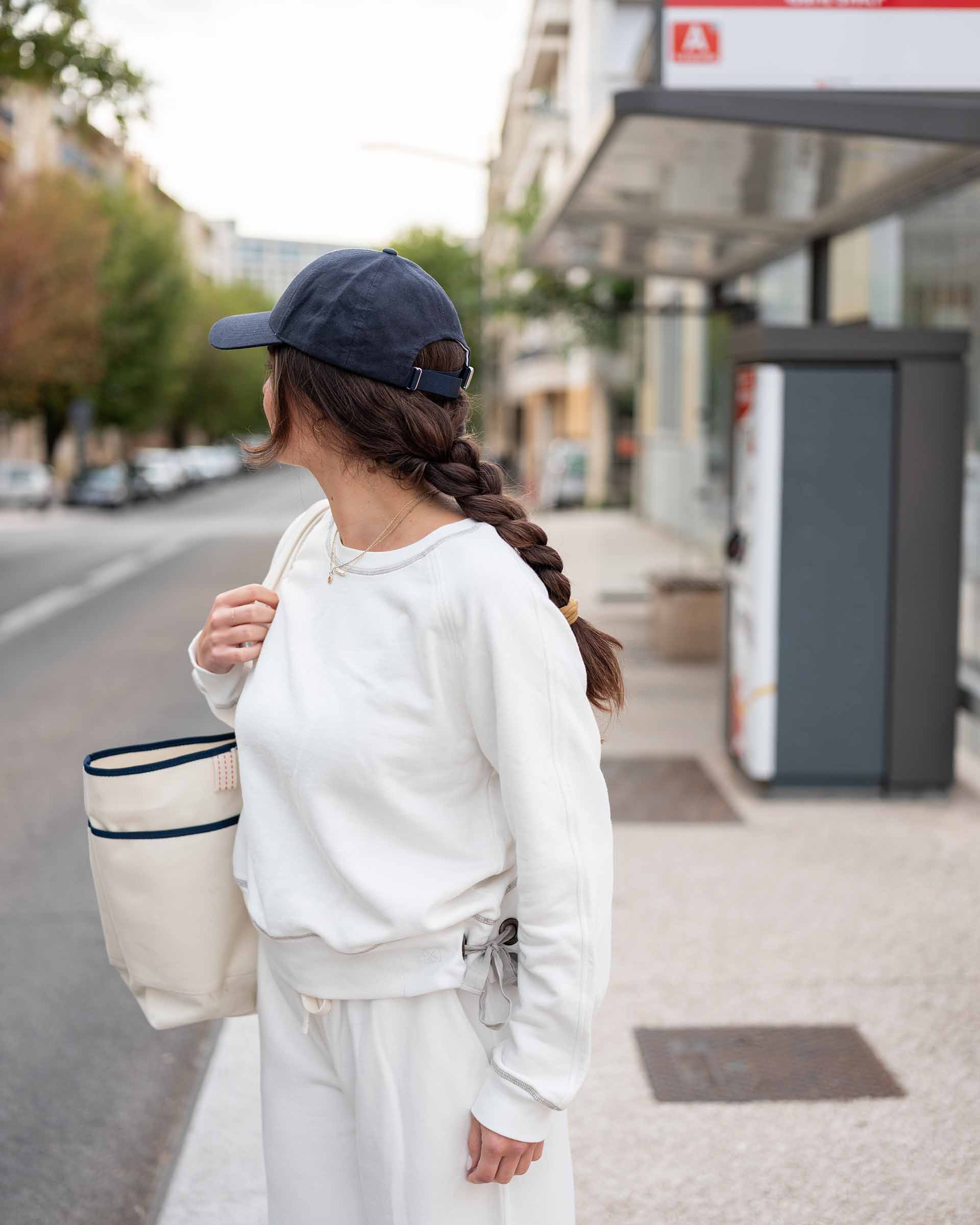 Person wearing a white sweatshirt and blue cap, holding a tote bag on a city street.