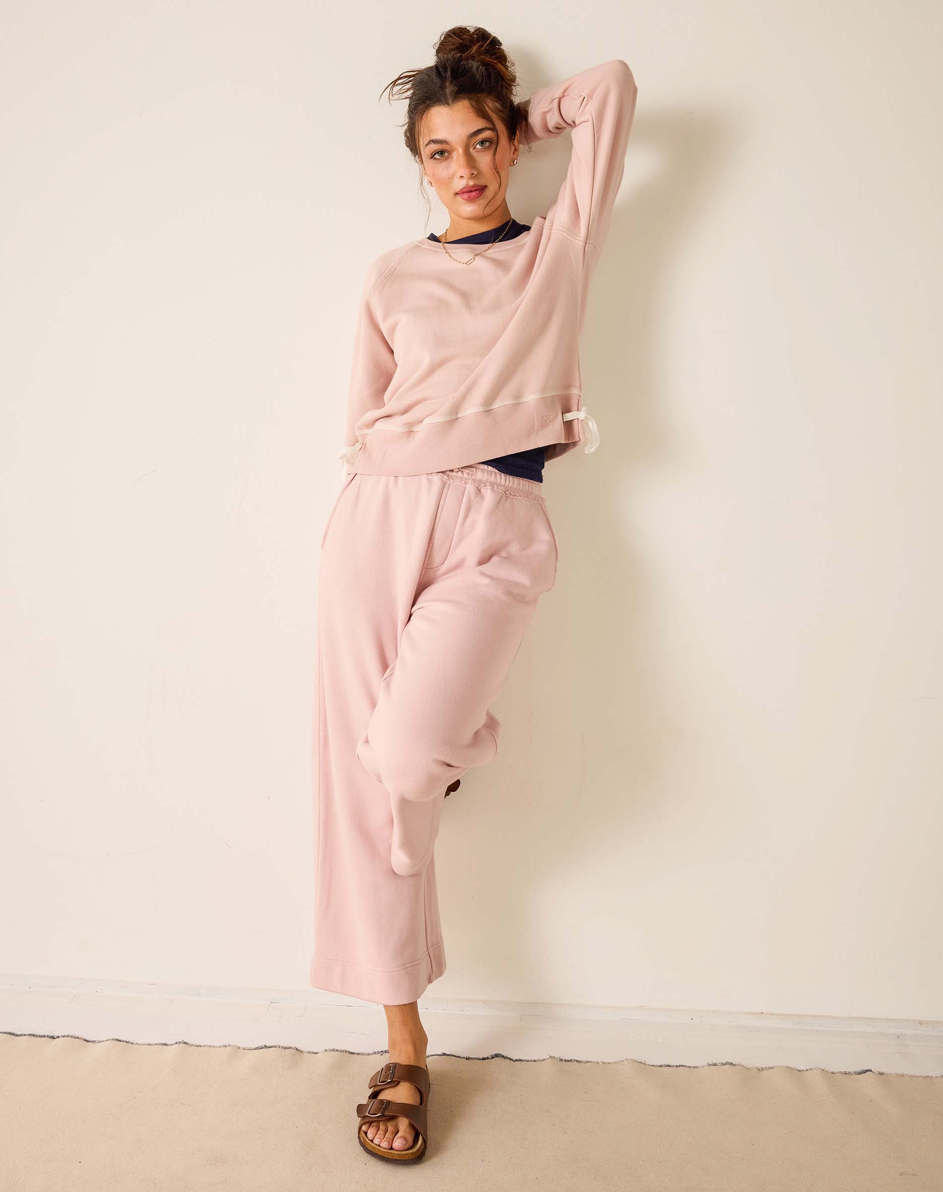 Woman wearing a pink sweatshirt and pants against a white wall.