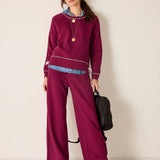 Woman wearing a maroon outfit with wide-leg pants and a sweater, holding a black bag against a plain background.