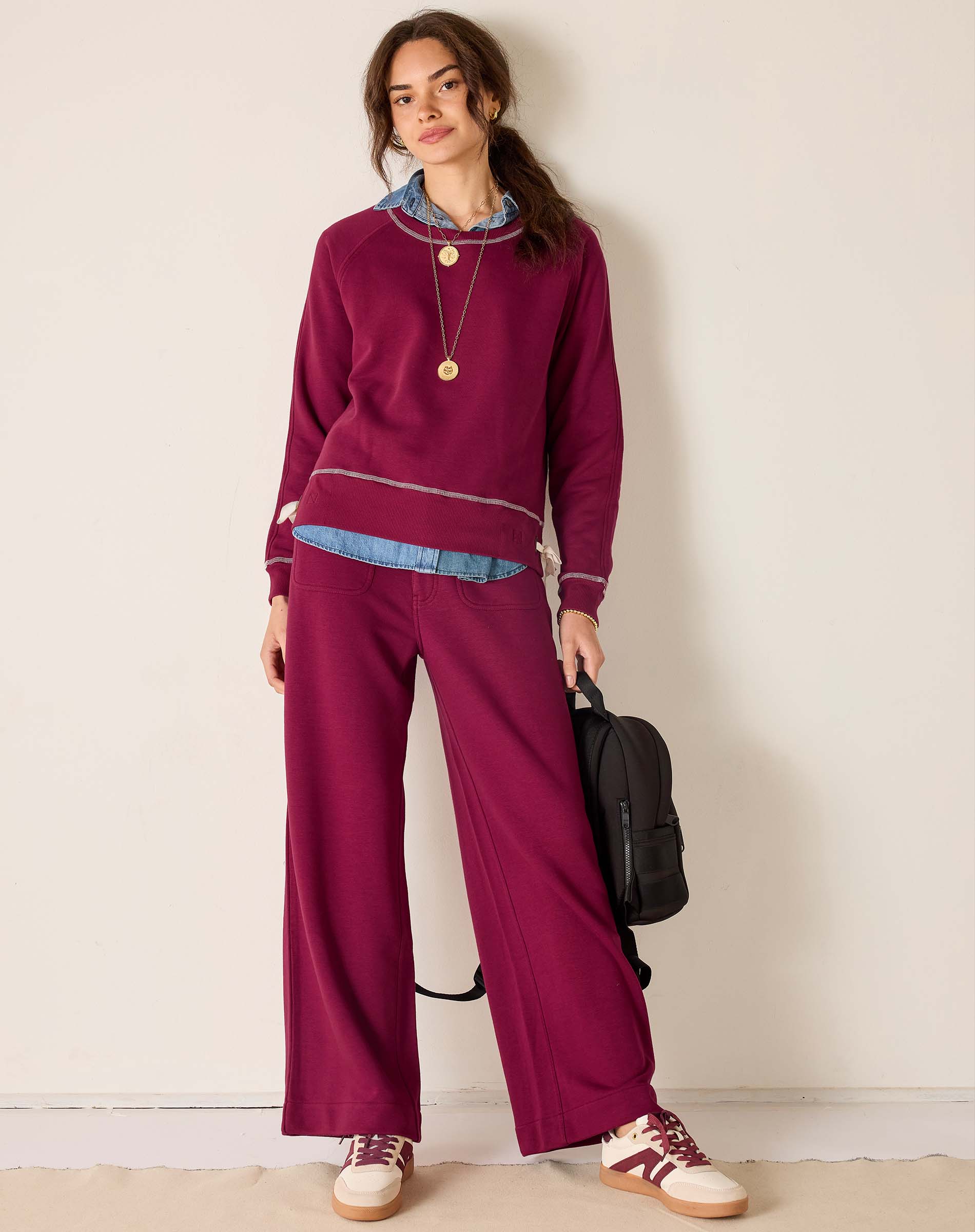 Woman wearing a maroon outfit with wide-leg pants and a sweater, holding a black bag against a plain background.