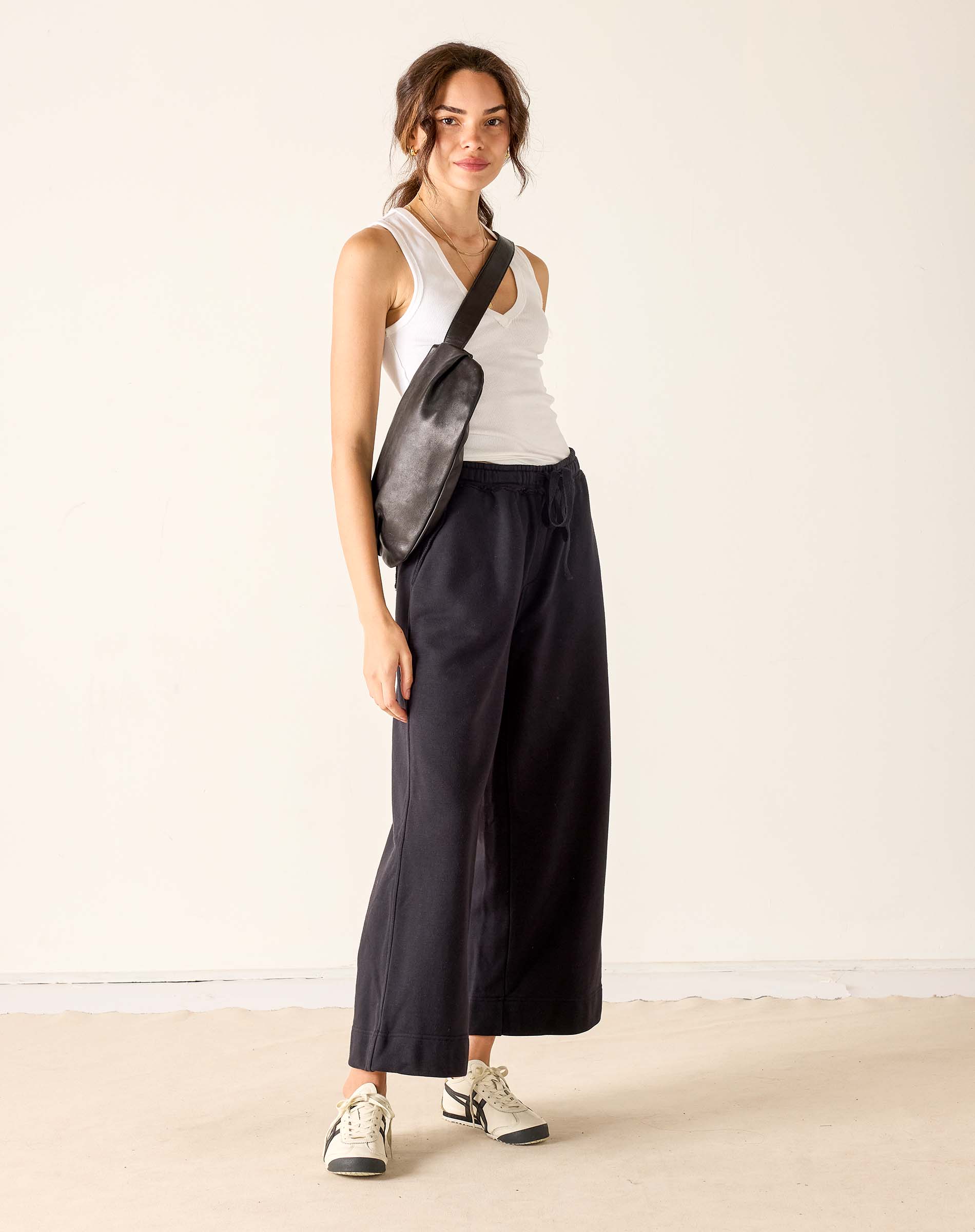 Woman wearing a white tank top, black wide-leg pants, and a black crossbody bag on a plain background