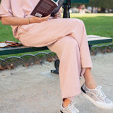 Person sitting on a bench in a park, wearing a pink outfit and white sneakers, holding a book.