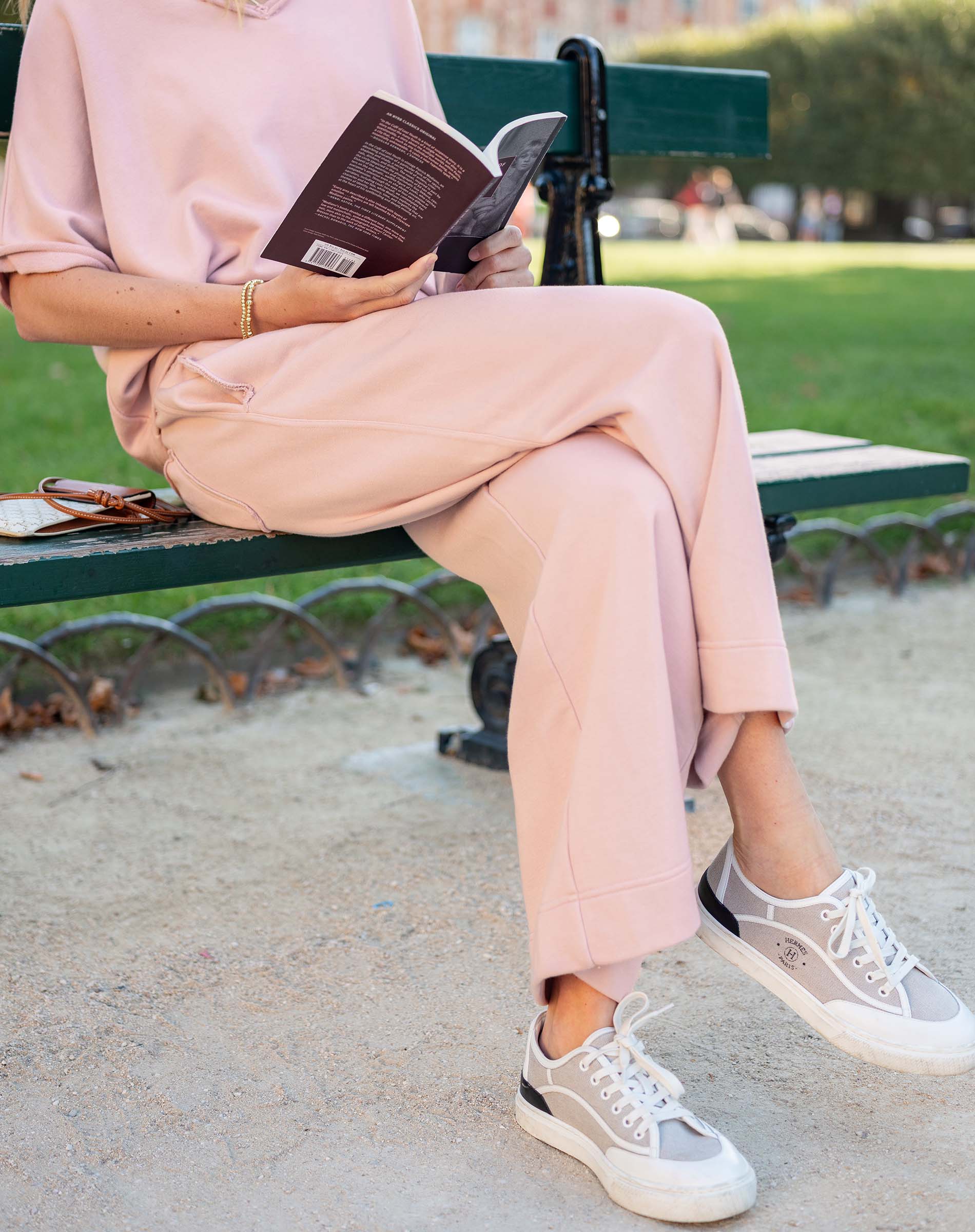 Person sitting on a bench in a park, wearing a pink outfit and white sneakers, holding a book.