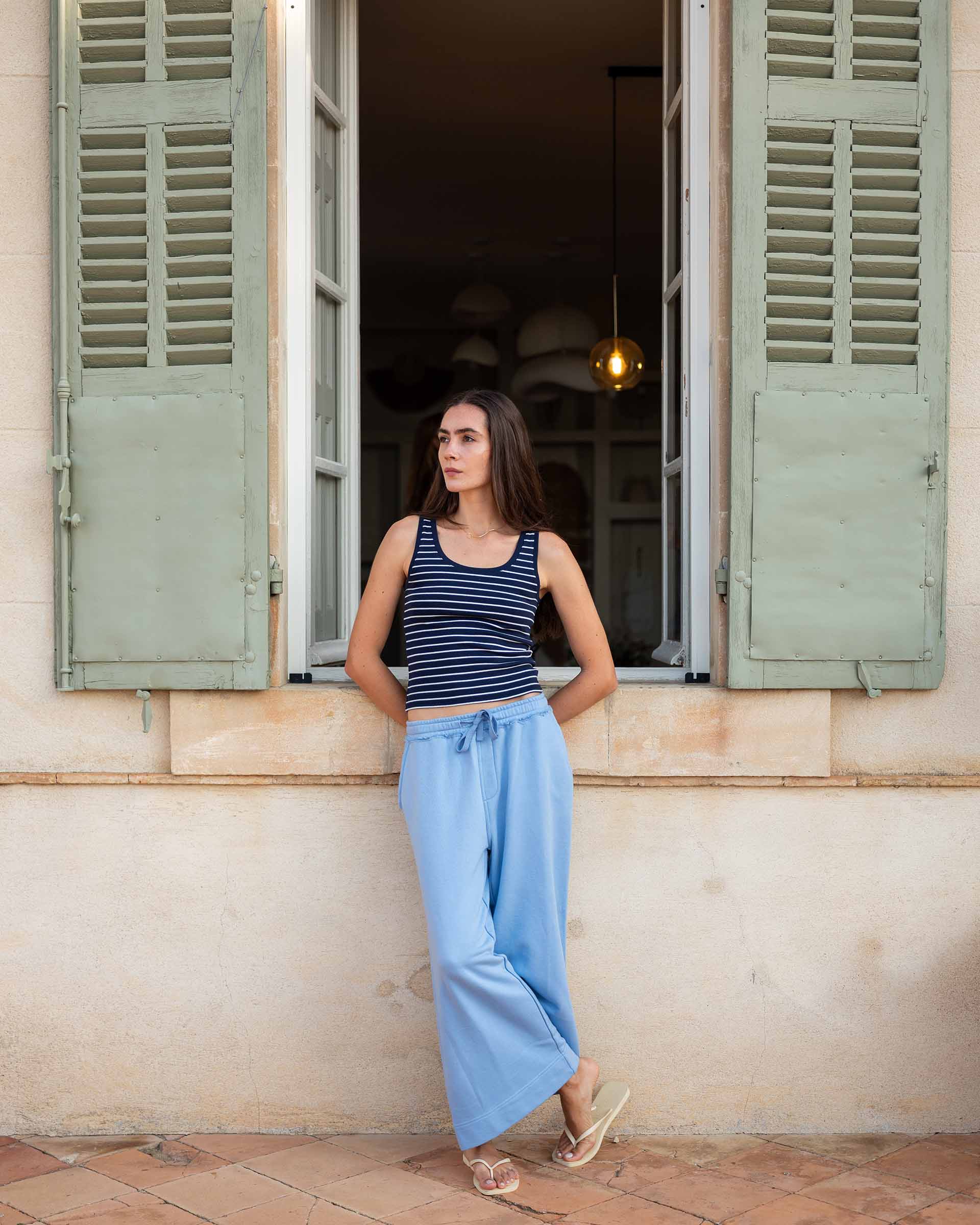 Woman standing in front of an open window with green shutters on a sunny day.