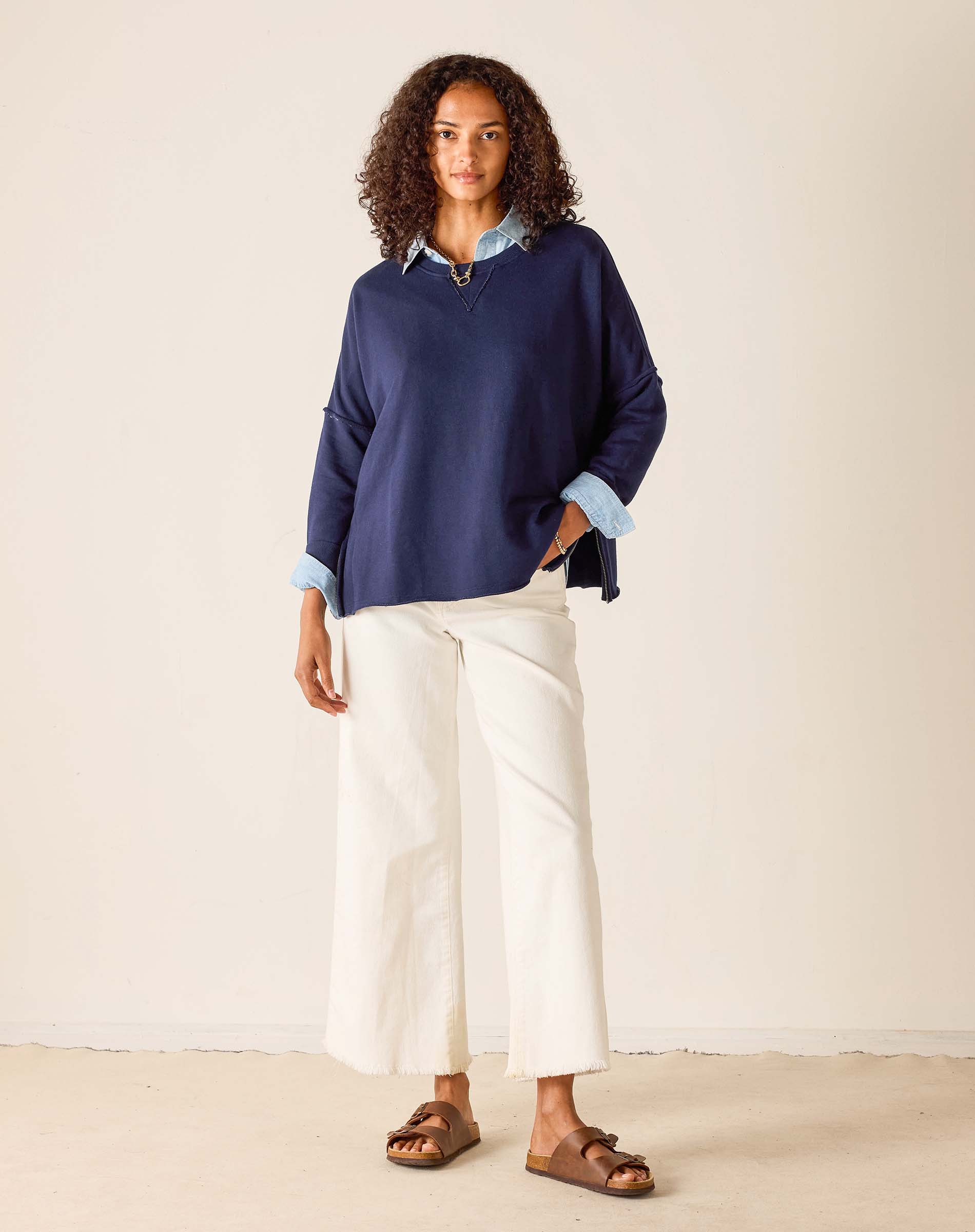 Woman wearing a navy sweater and white pants against a beige background