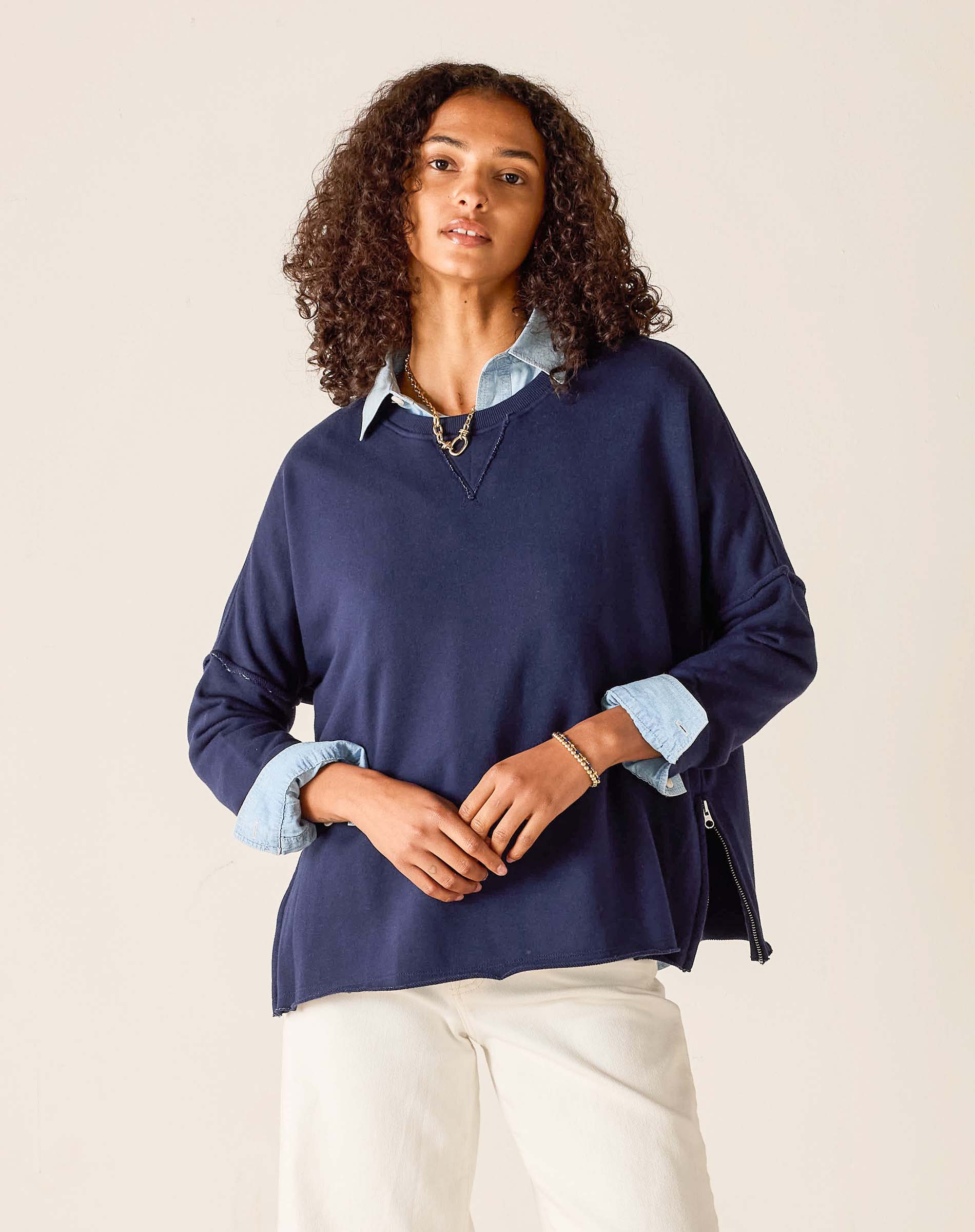 Woman wearing a navy blue sweater over a light blue shirt with white pants on a beige background