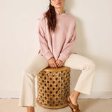 Woman sitting on a woven stool wearing a pink sweatshirt and white pants.