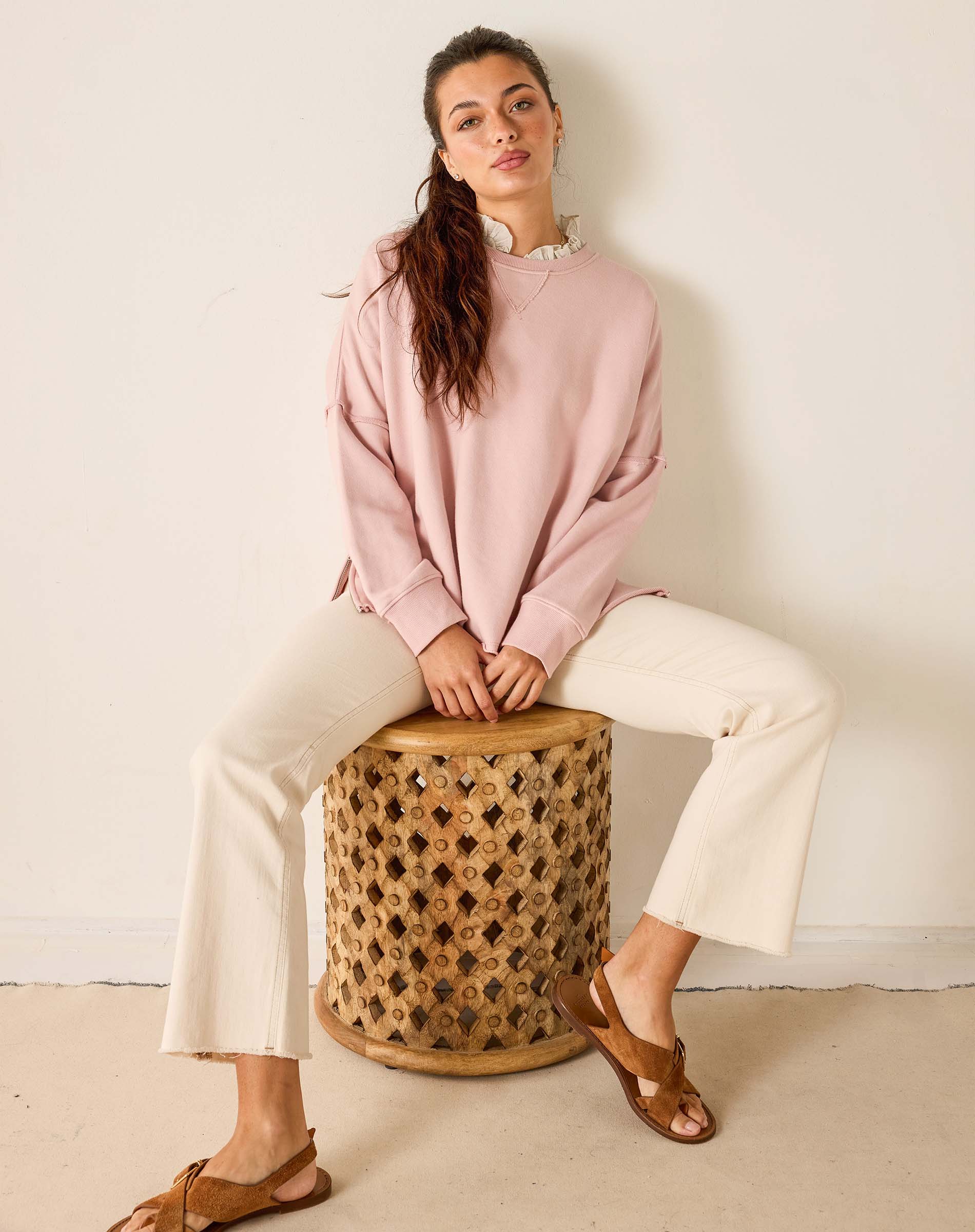 Woman sitting on a woven stool wearing a pink sweatshirt and white pants.