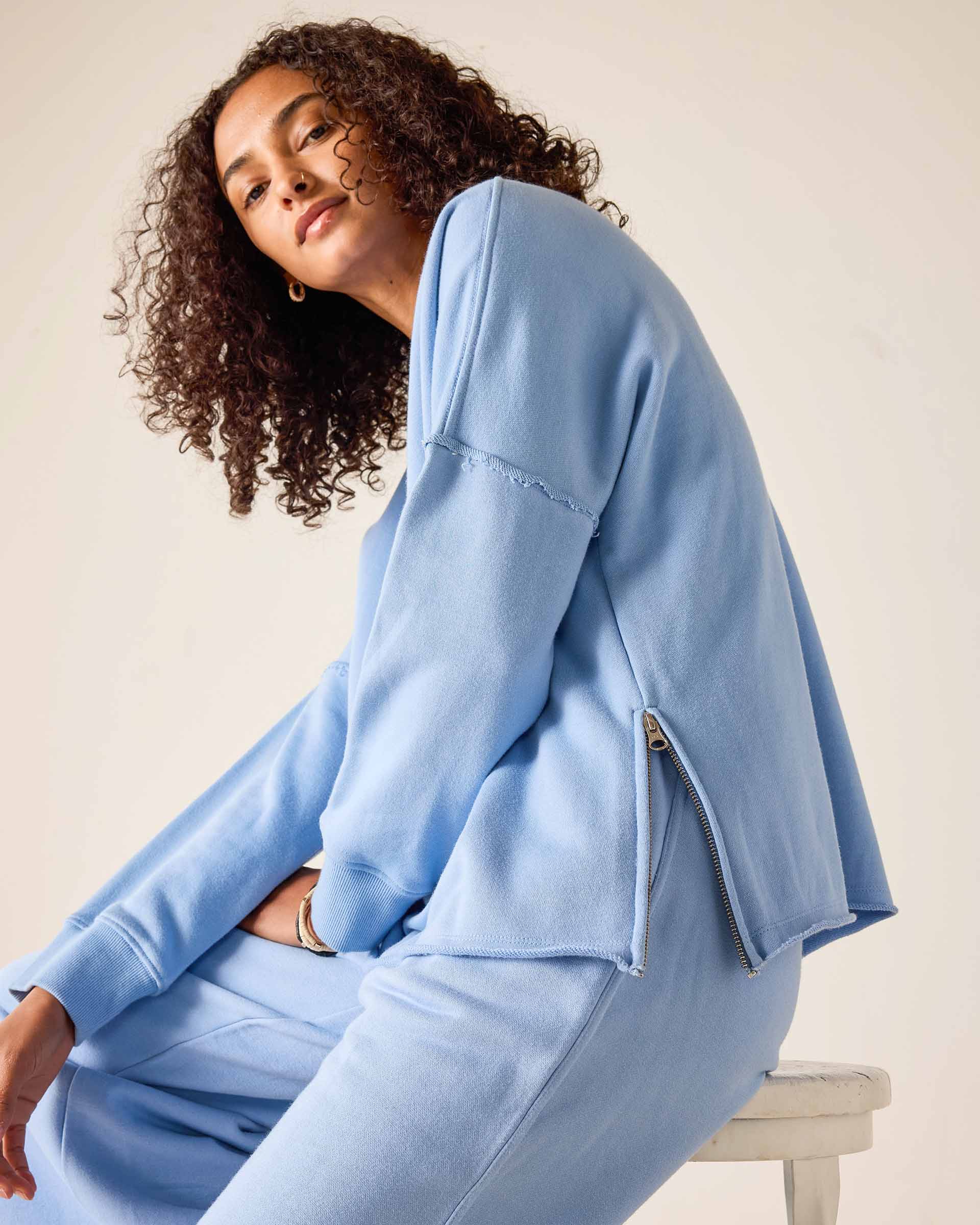 Woman wearing a light blue tracksuit sitting on a white stool against a beige background