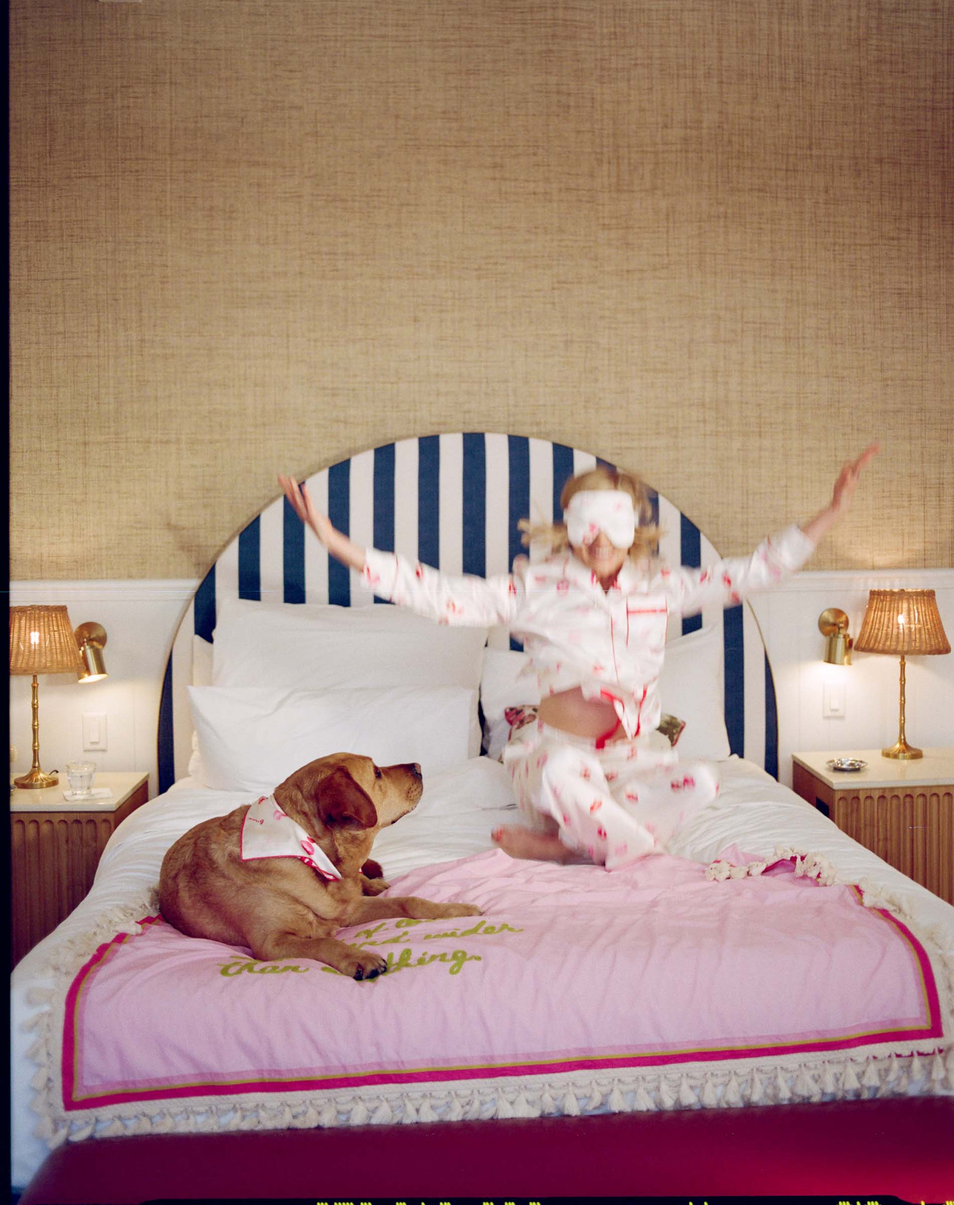 Woman in pajamas sitting on a bed with a dog, wearing a face mask, in a room with striped headboard and lamps.