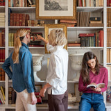 Three women in cozy knitwear and casual dresses share a moment together while browsing a bookshelf filled with books. One woman leans in to show something on a shelf, another reads a book intently, and the third looks on from the side.