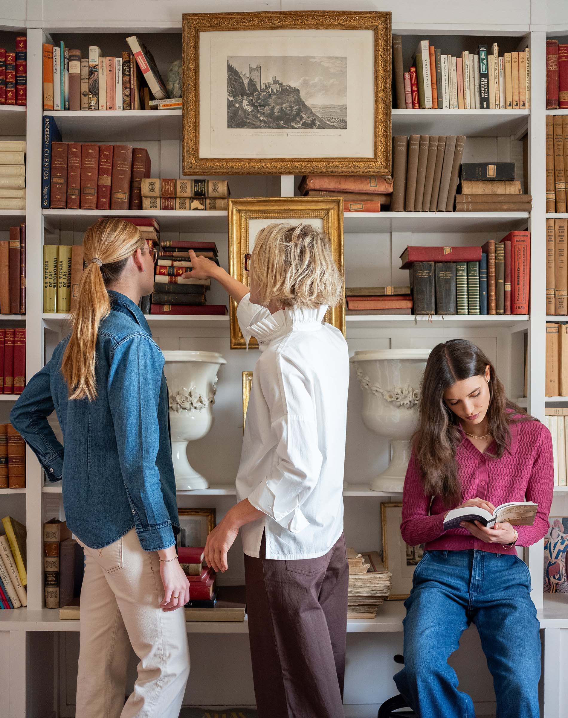 Three women in cozy knitwear and casual dresses share a moment together while browsing a bookshelf filled with books. One woman leans in to show something on a shelf, another reads a book intently, and the third looks on from the side.