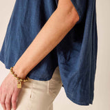 Person wearing a blue denim shirt and beige pants with a gold bracelet on a neutral background