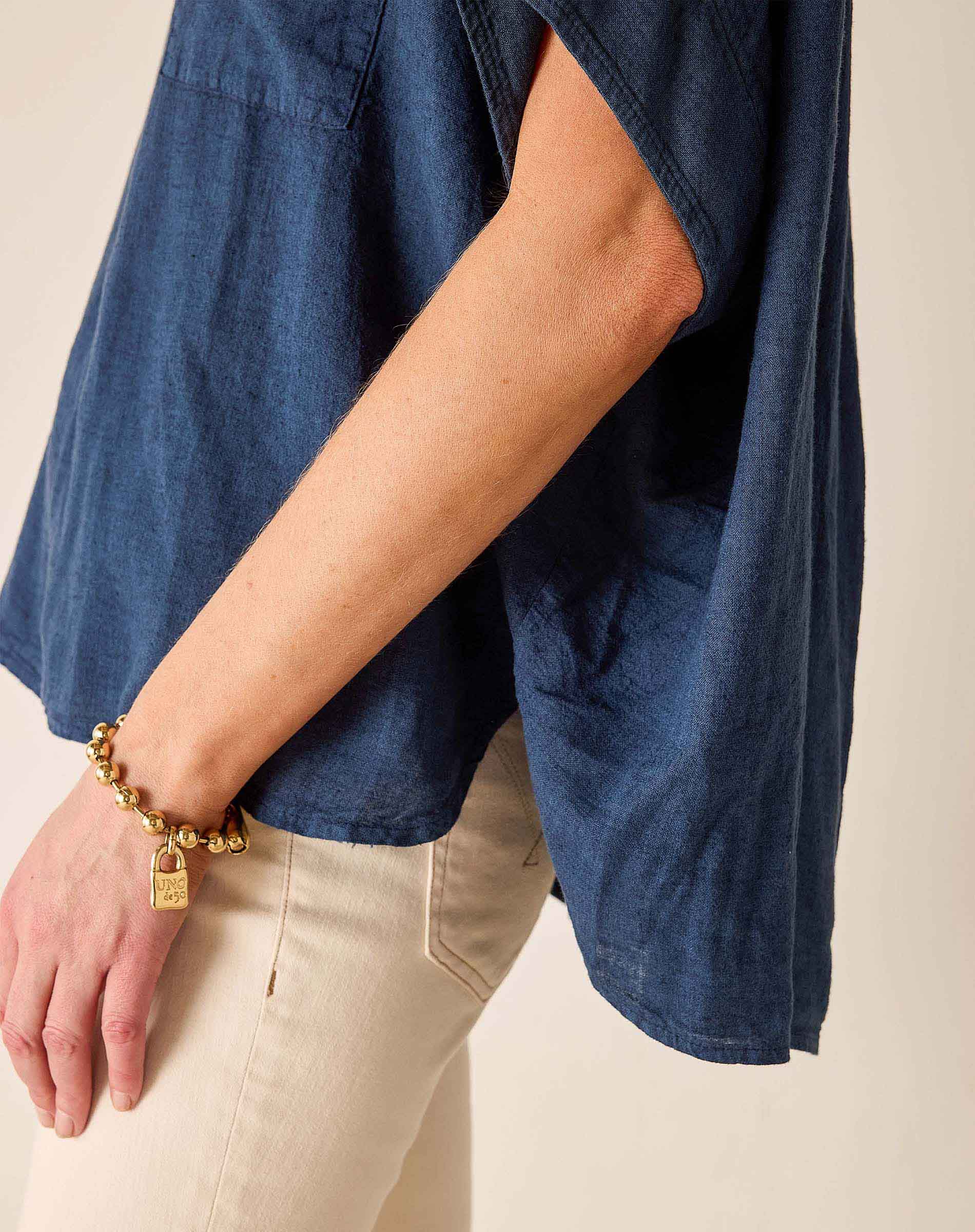 Person wearing a blue denim shirt and beige pants with a gold bracelet on a neutral background