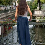 Woman in a blue skirt and striped top standing by a canal with greenery and buildings in the background.