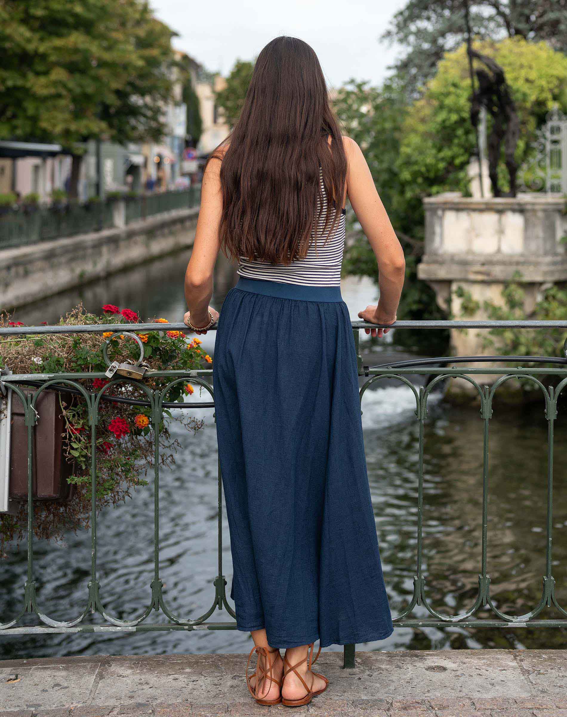 Woman in a blue skirt and striped top standing by a canal with greenery and buildings in the background.