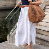 Woman in a navy top and white skirt holding a brown woven bag on steps by water.