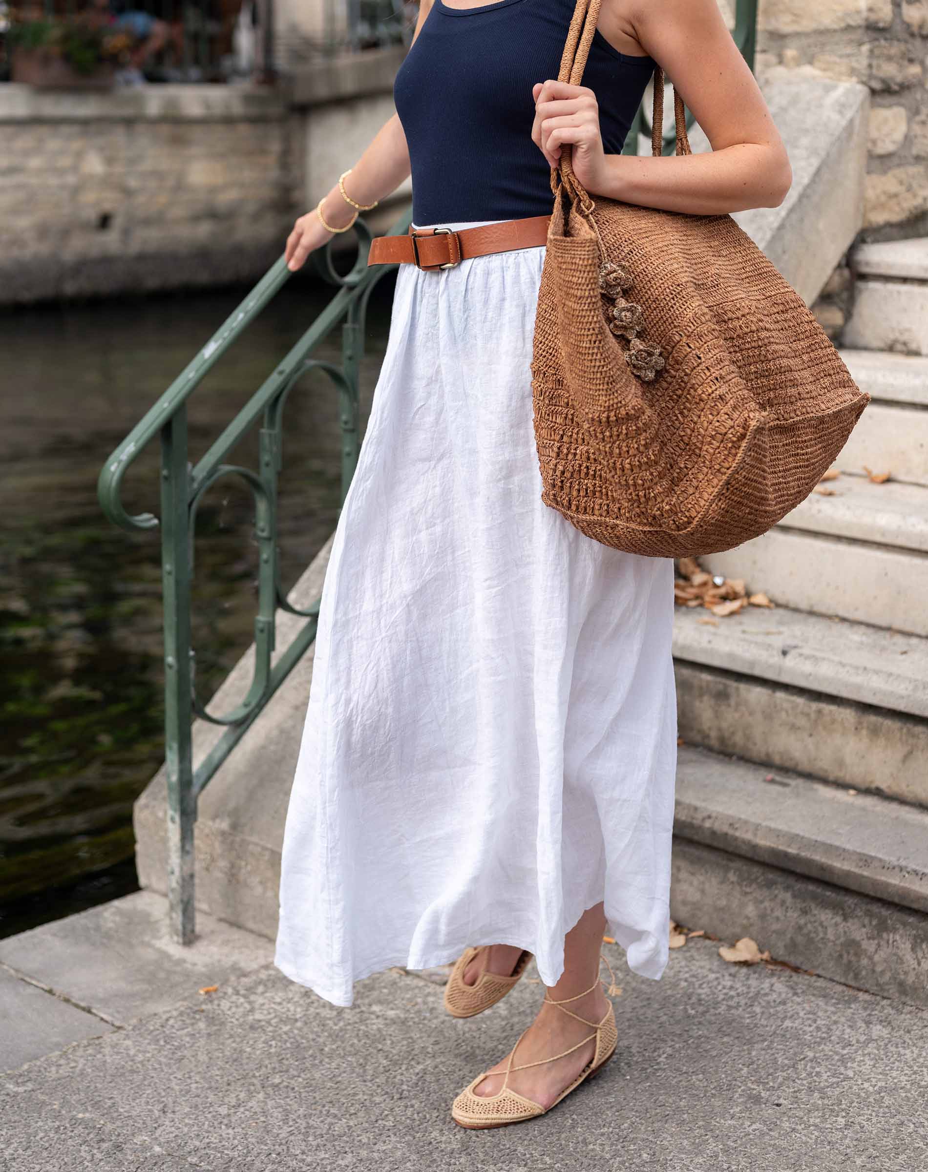 Woman in a navy top and white skirt holding a brown woven bag on steps by water.