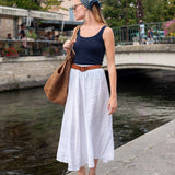 Woman in a navy tank top and white skirt standing by a waterfront with a brown bag.