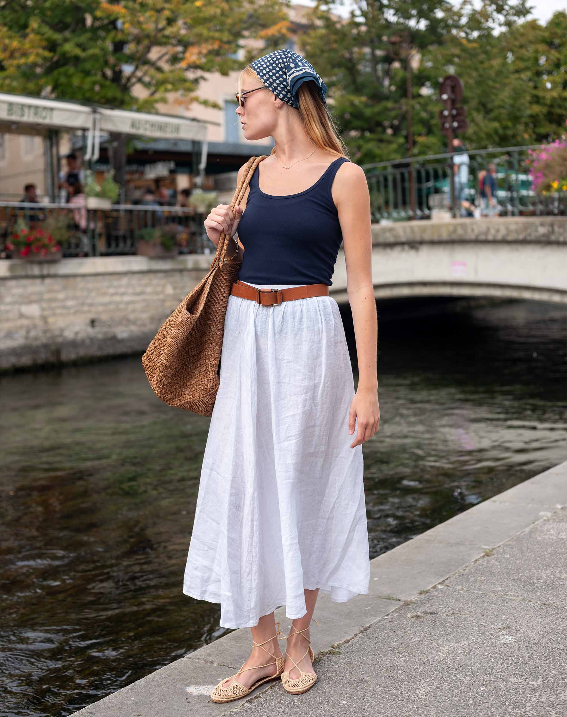 Woman in a navy tank top and white skirt standing by a waterfront with a brown bag.