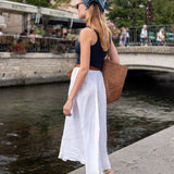 Woman in a black top and white pants standing by a waterfront with a brown bag.
