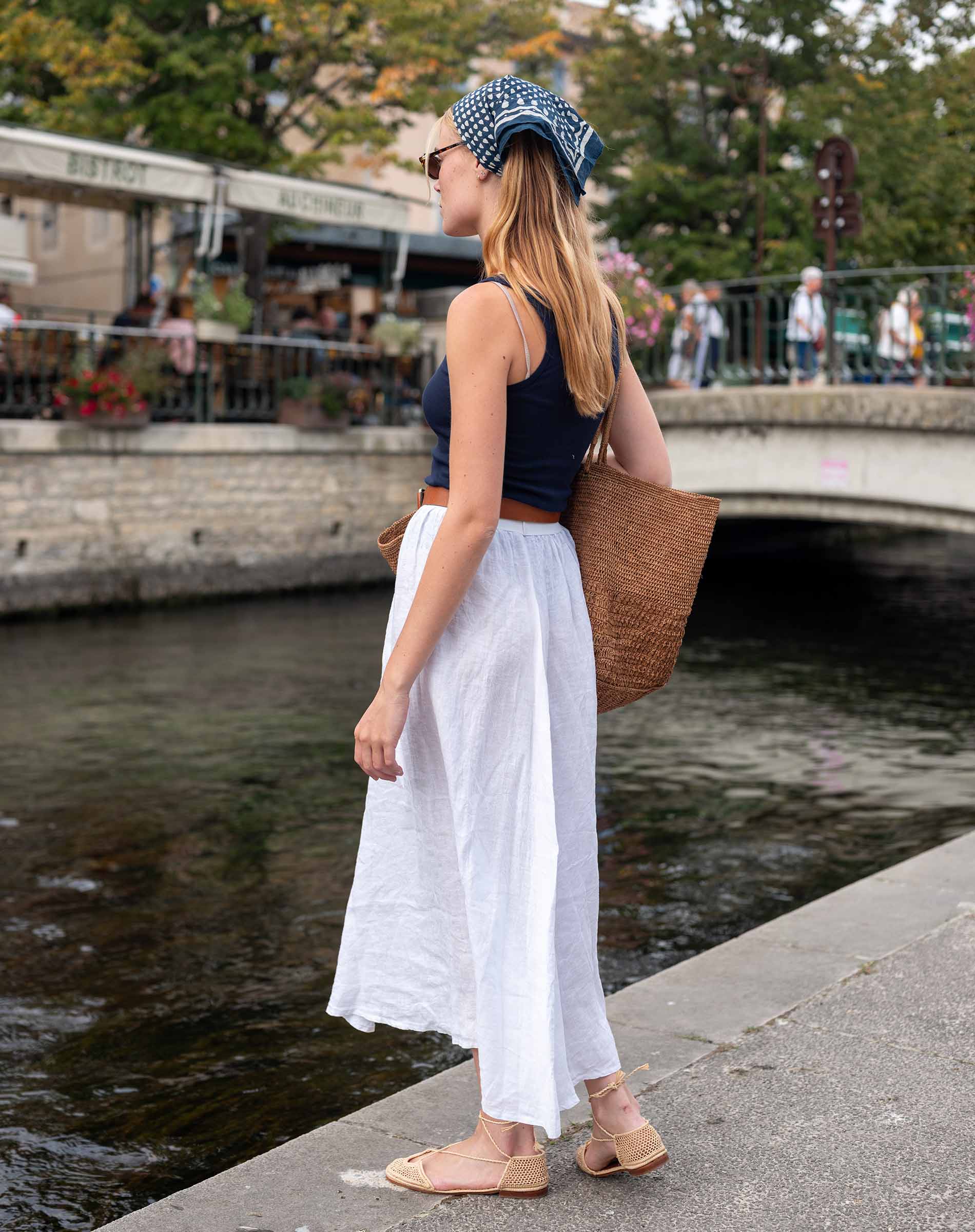 Woman in a black top and white pants standing by a waterfront with a brown bag.