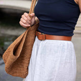 Person wearing a navy tank top, white pants, and brown belt holding a woven bag.