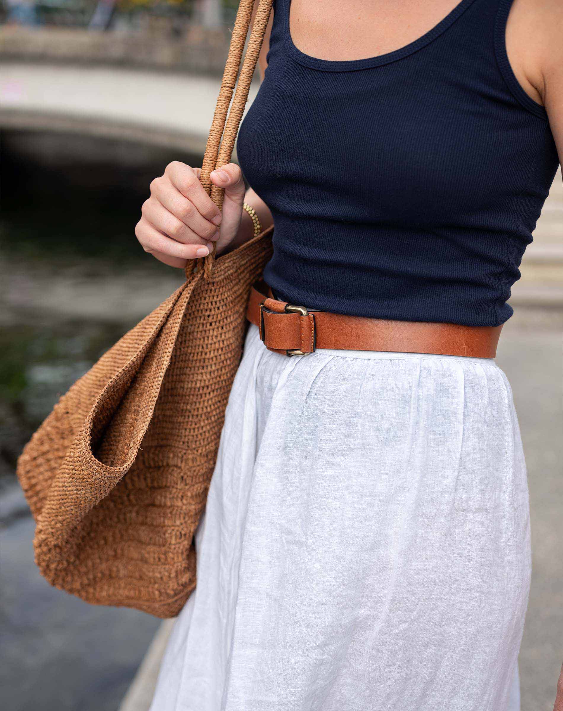 Person wearing a navy tank top, white pants, and brown belt holding a woven bag.