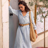 Woman in a light blue dress with a brown belt standing next to a door, holding a woven bag.