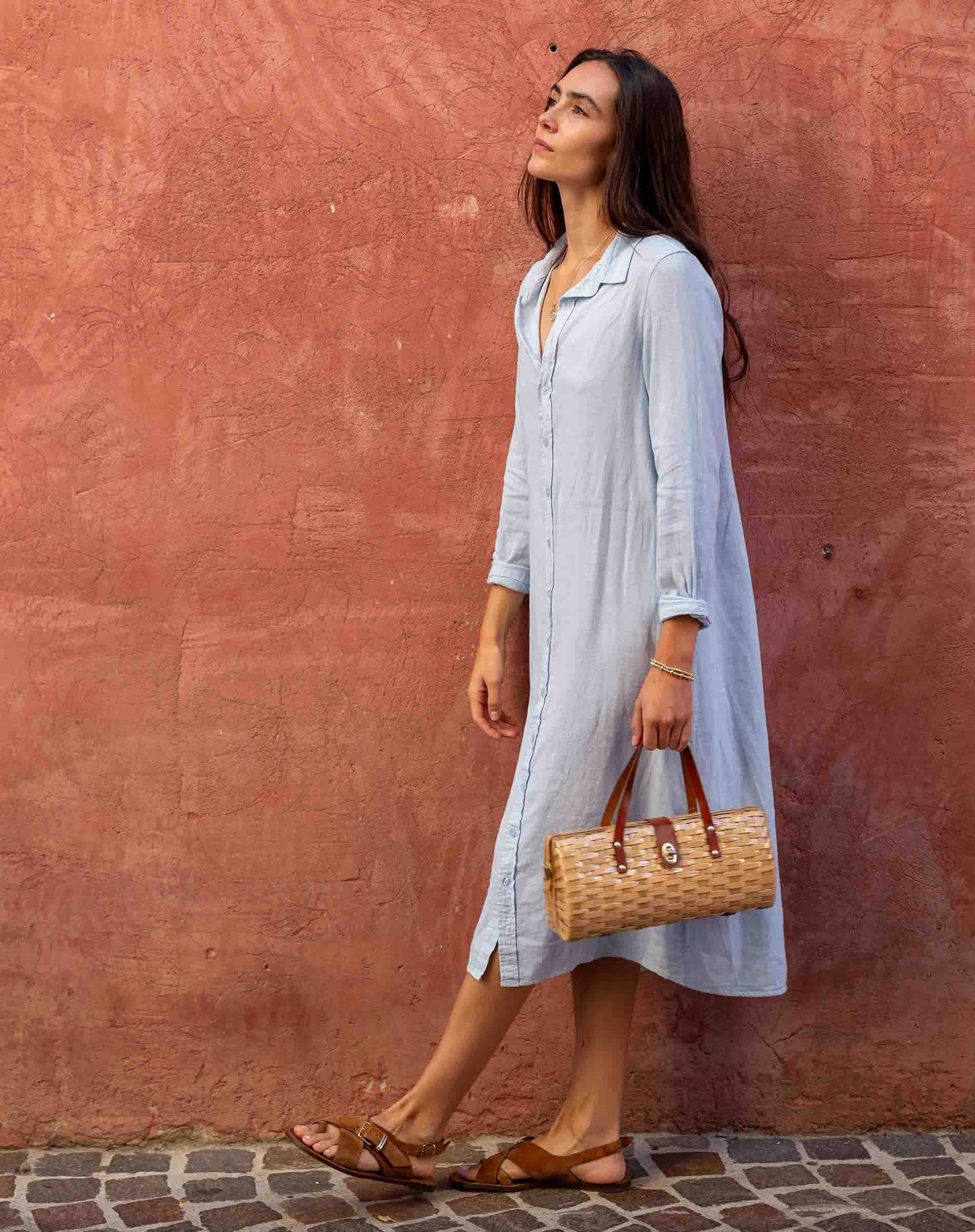 Woman in a light blue dress holding a woven bag against a red wall.