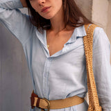 Woman wearing a light blue shirt with a brown belt and holding a beige woven bag.