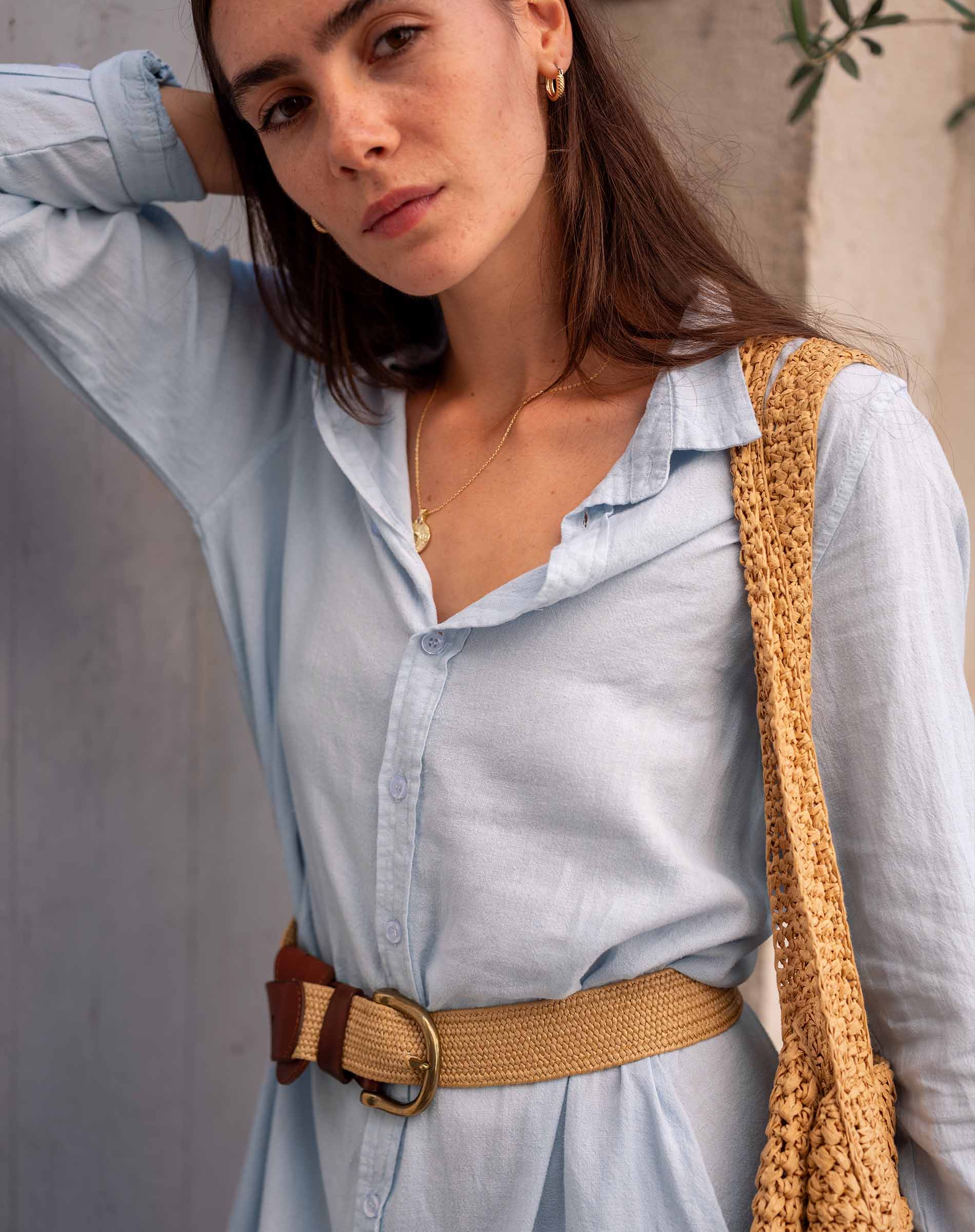 Woman wearing a light blue shirt with a brown belt and holding a beige woven bag.