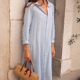 Woman wearing a light blue dress holding a woven bag against a stone wall.