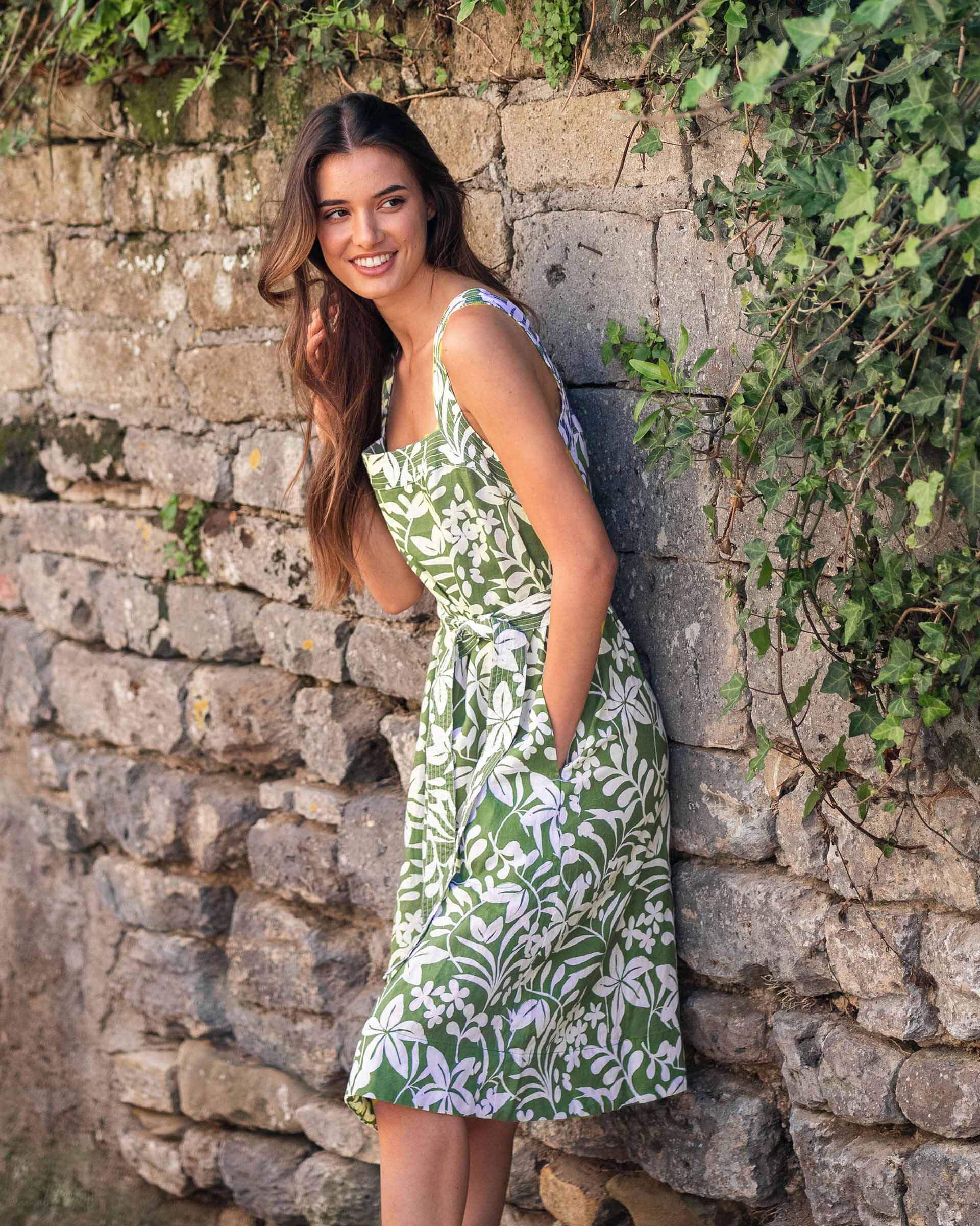 female wearing a green linen dress with white floral print and a self belt leaning on a stone wall