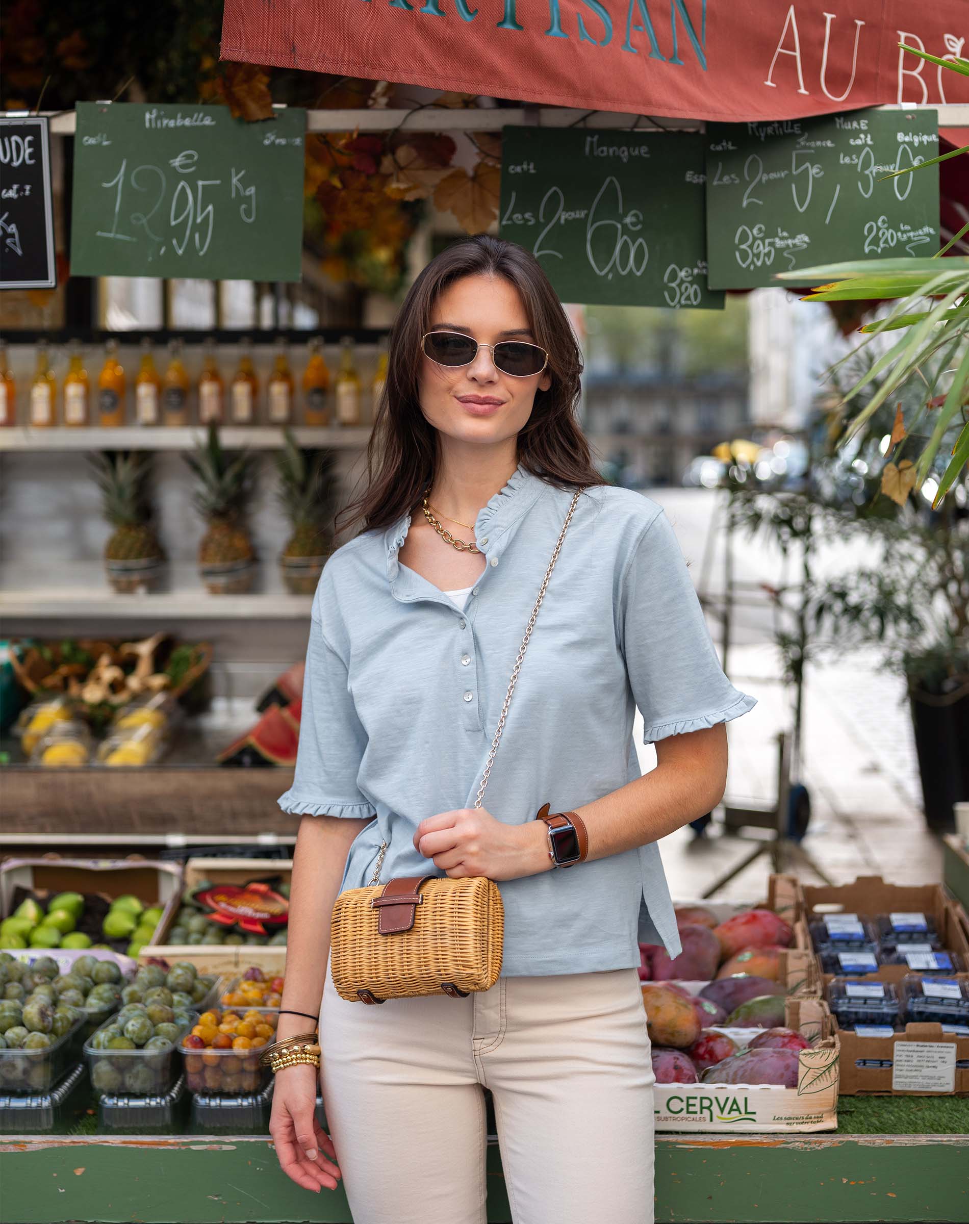 Woman standing in front of a market stall with produce and signs.