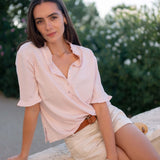 Woman in a light pink shirt and white shorts sitting outdoors with greenery in the background