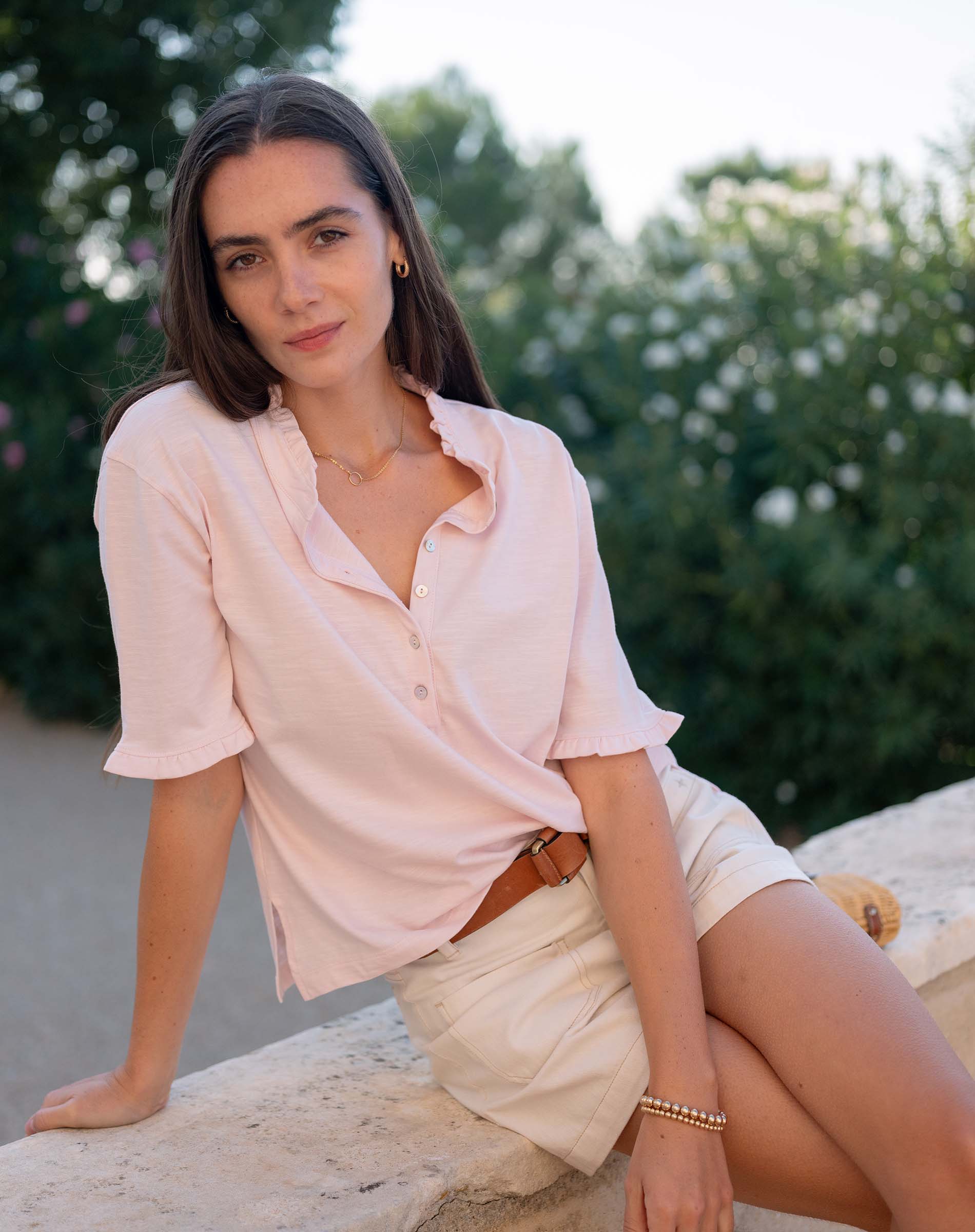 Woman in a light pink shirt and white shorts sitting outdoors with greenery in the background