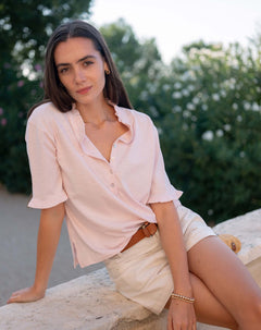 Woman in a light pink shirt and white shorts sitting outdoors with greenery in the background