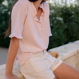 Woman wearing a light pink blouse and beige shorts sitting outdoors.