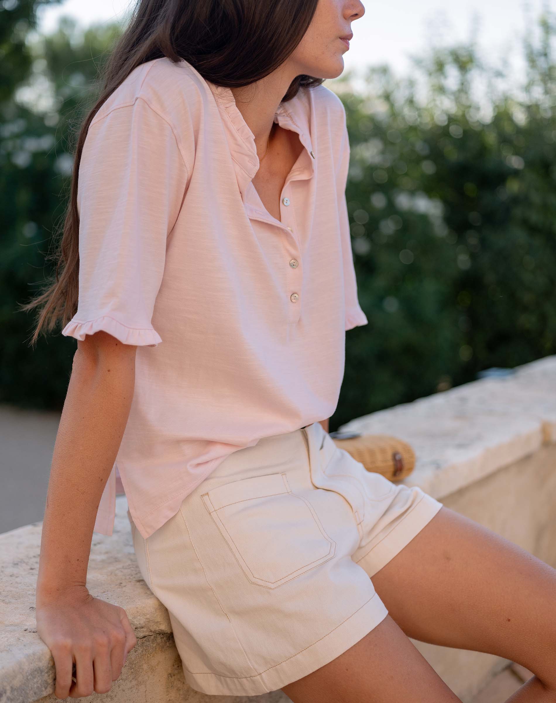 Woman wearing a light pink blouse and beige shorts sitting outdoors.