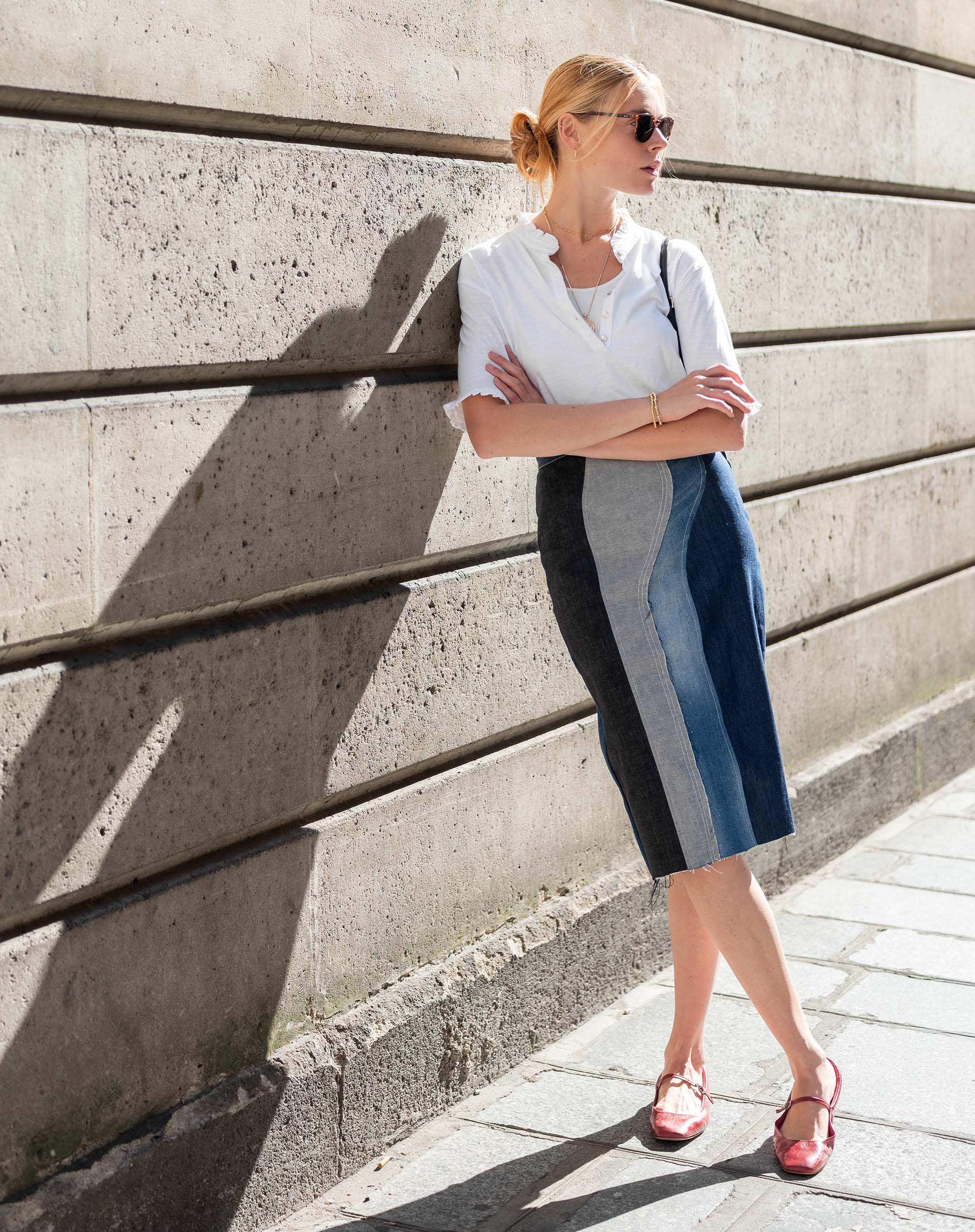Woman in a white blouse and blue skirt standing against a concrete wall.