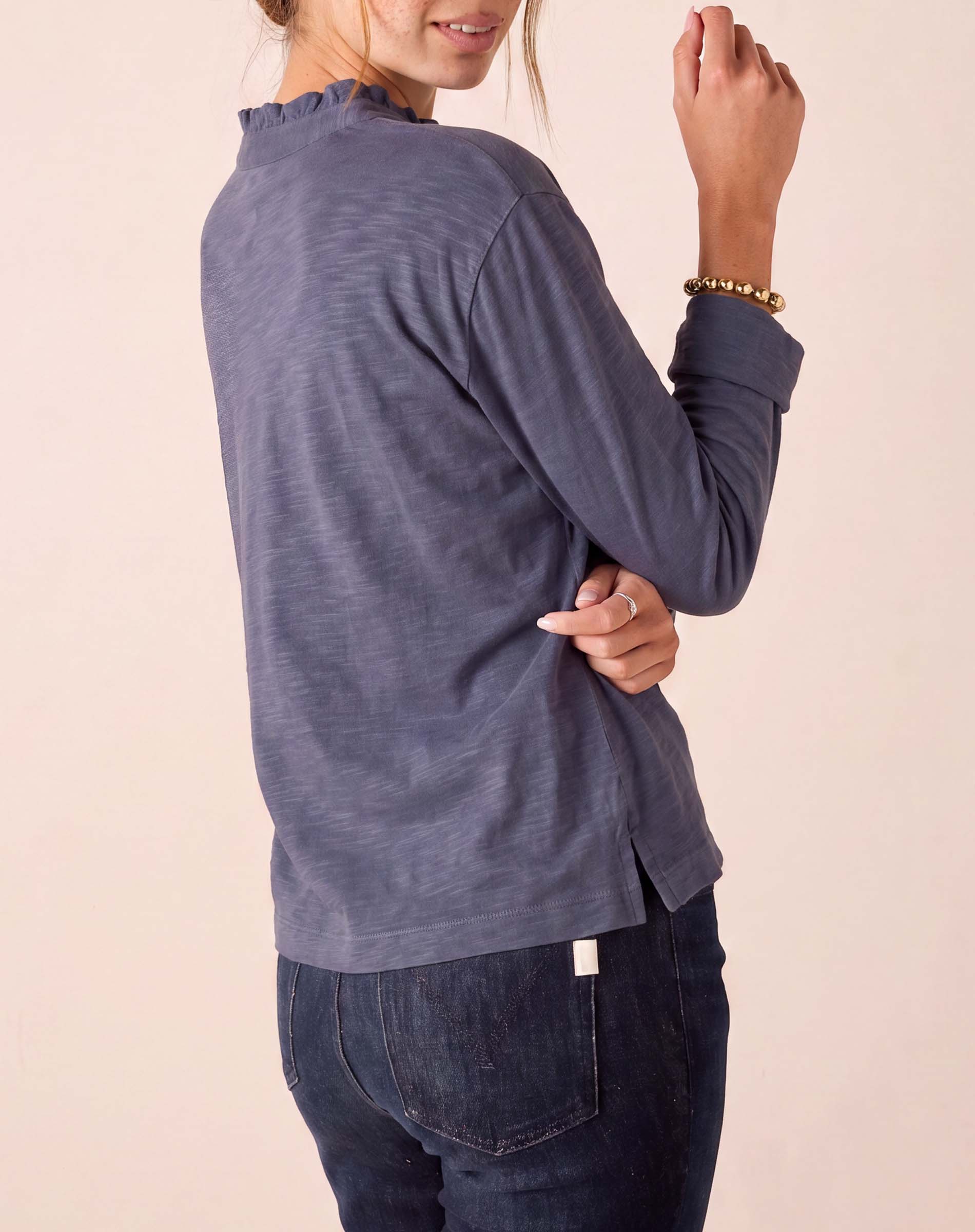 Side view of the woman wearing a women's travel-friendly dusty blue long-sleeve ruffle henley top and dark denim jeans.
