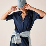 Person wearing a navy blue shirt and pants with a denim cap and belt against a beige background