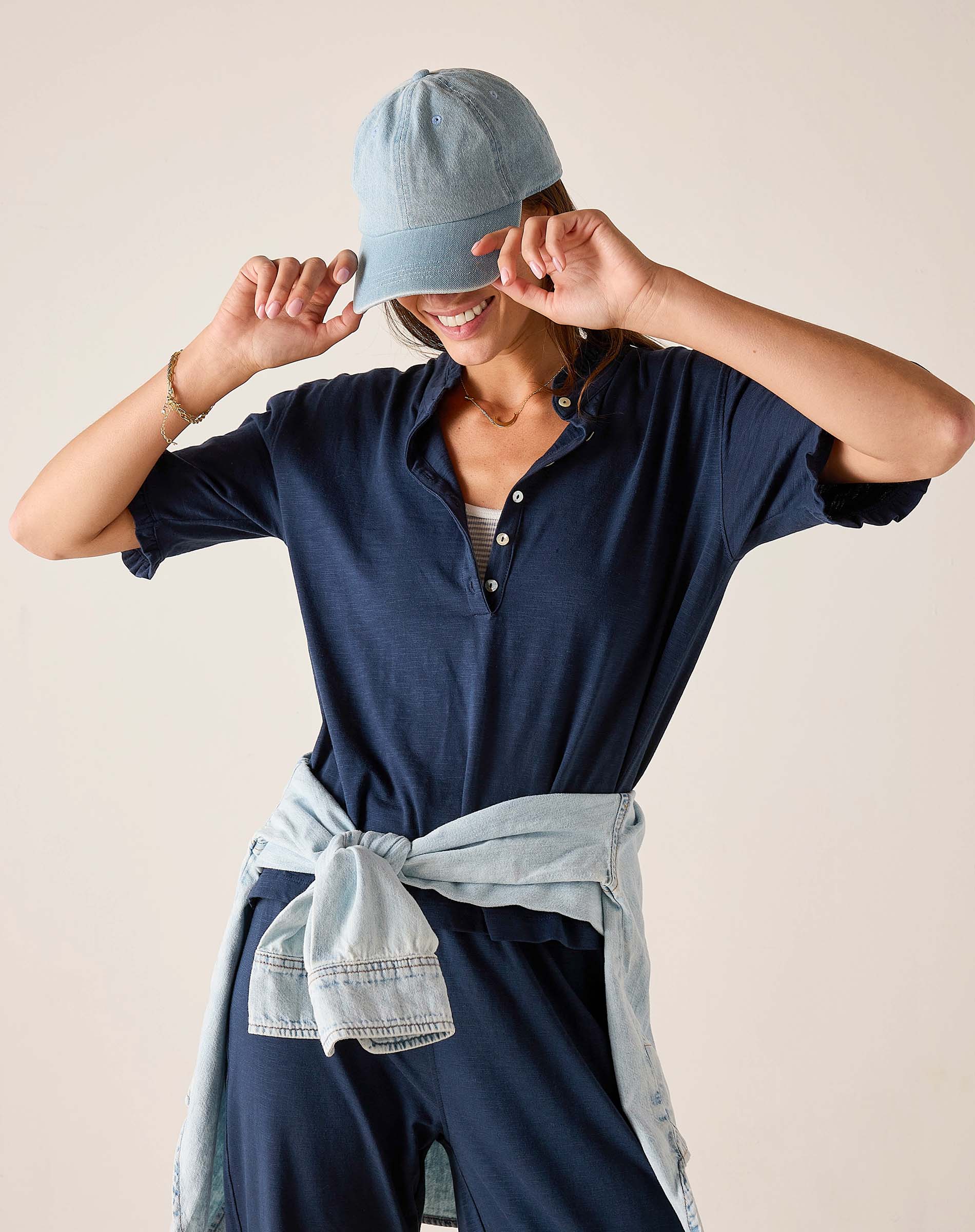Person wearing a navy blue shirt and pants with a denim cap and belt against a beige background