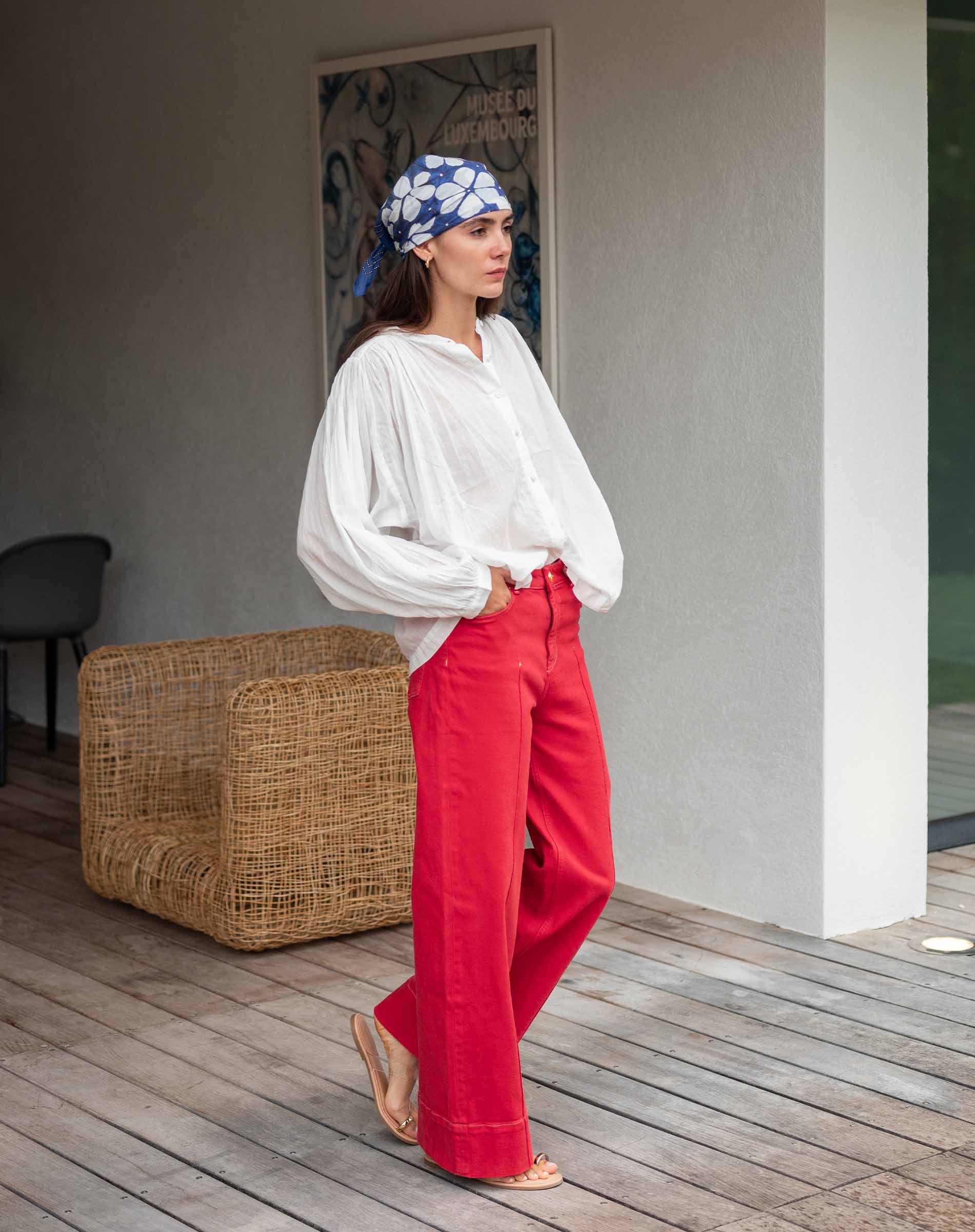 Woman wearing a white blouse and red pants with a blue patterned headscarf, standing on a wooden deck.