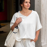 Woman in a white outfit holding a beige tote bag, standing against a neutral wall.