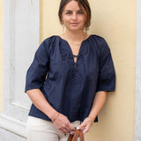 Woman wearing a navy blue blouse and white pants, standing against a light-colored wall.