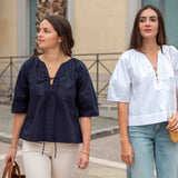 Two women standing outdoors, one in a navy blue top and beige pants, the other in a white top and light blue jeans.
