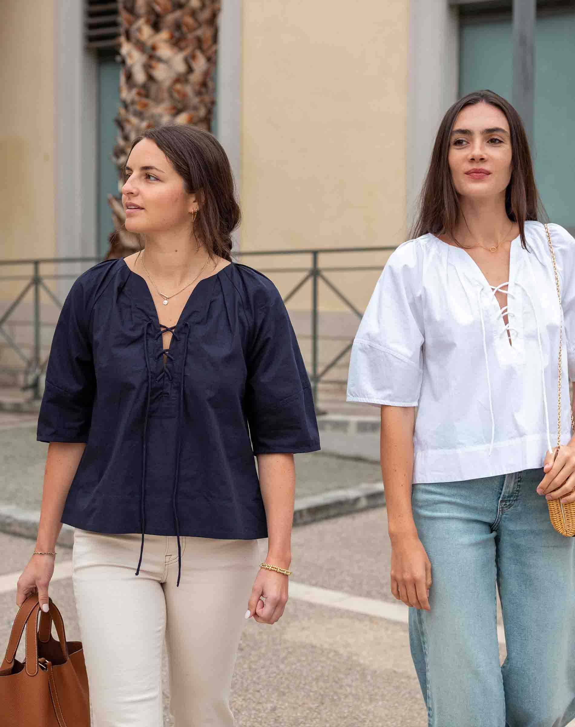 Two women standing outdoors, one in a navy blue top and beige pants, the other in a white top and light blue jeans.